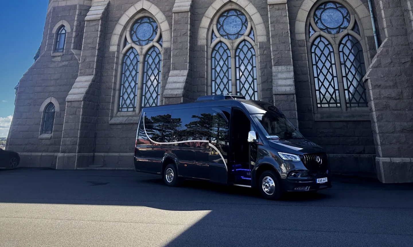 Öckeröbussarnas VIP-buss, en svart Mercedes Sprinter med öppen passagerardörr, framför en stenkyrka.